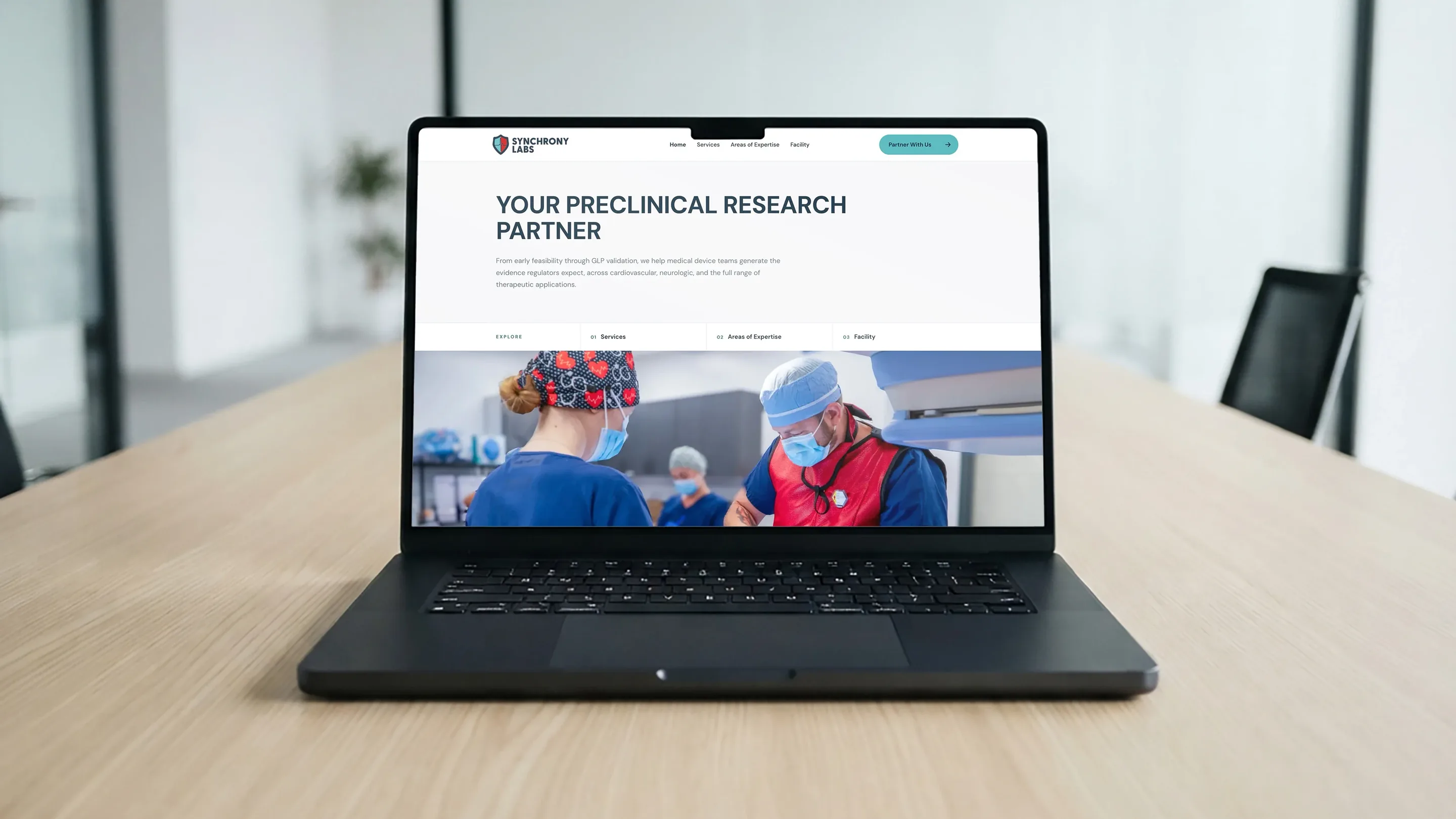 Synchrony Labs website open on a laptop on a conference table, its homepage headline reading Your Preclinical Research Partner