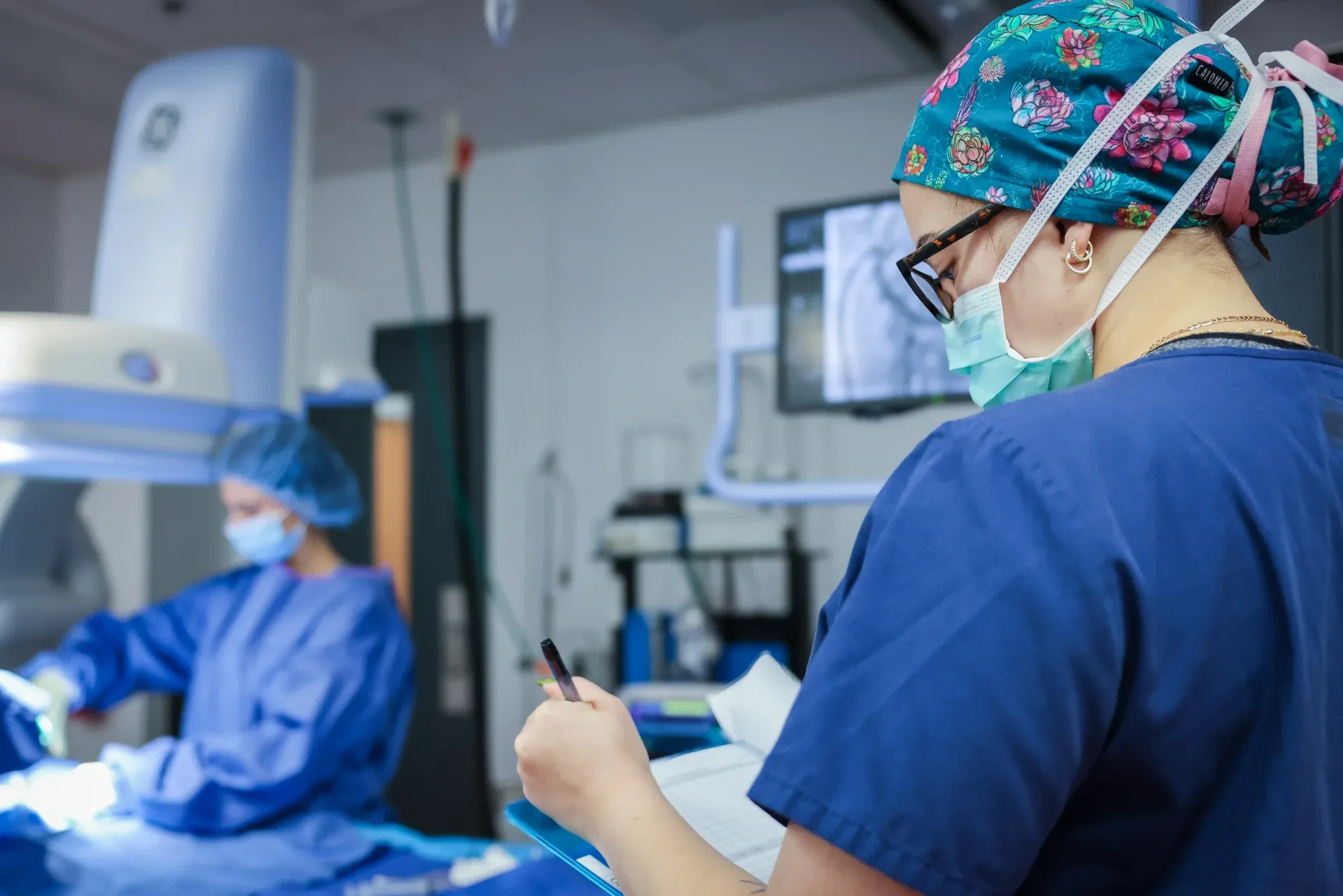 Synchrony Labs clinician in a floral scrub cap reviewing study notes beside an imaging monitor during a procedure